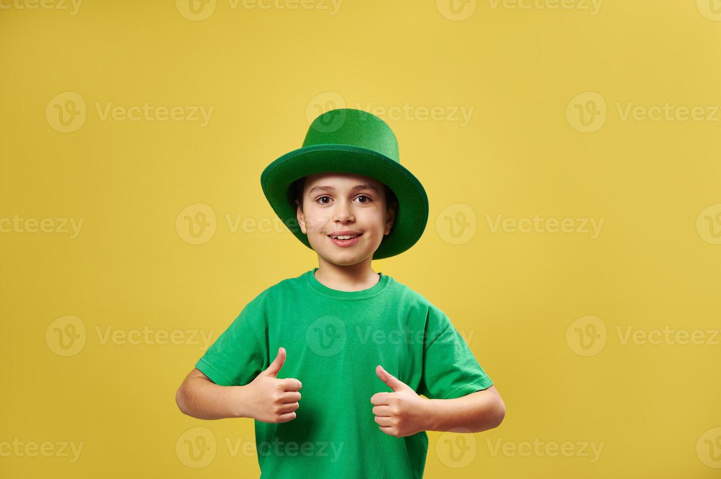 Smiling little boy in Irish green leprechaun hat showing thumbs up at camera standing on yellow background with copy space. Saint Patrick Day photo