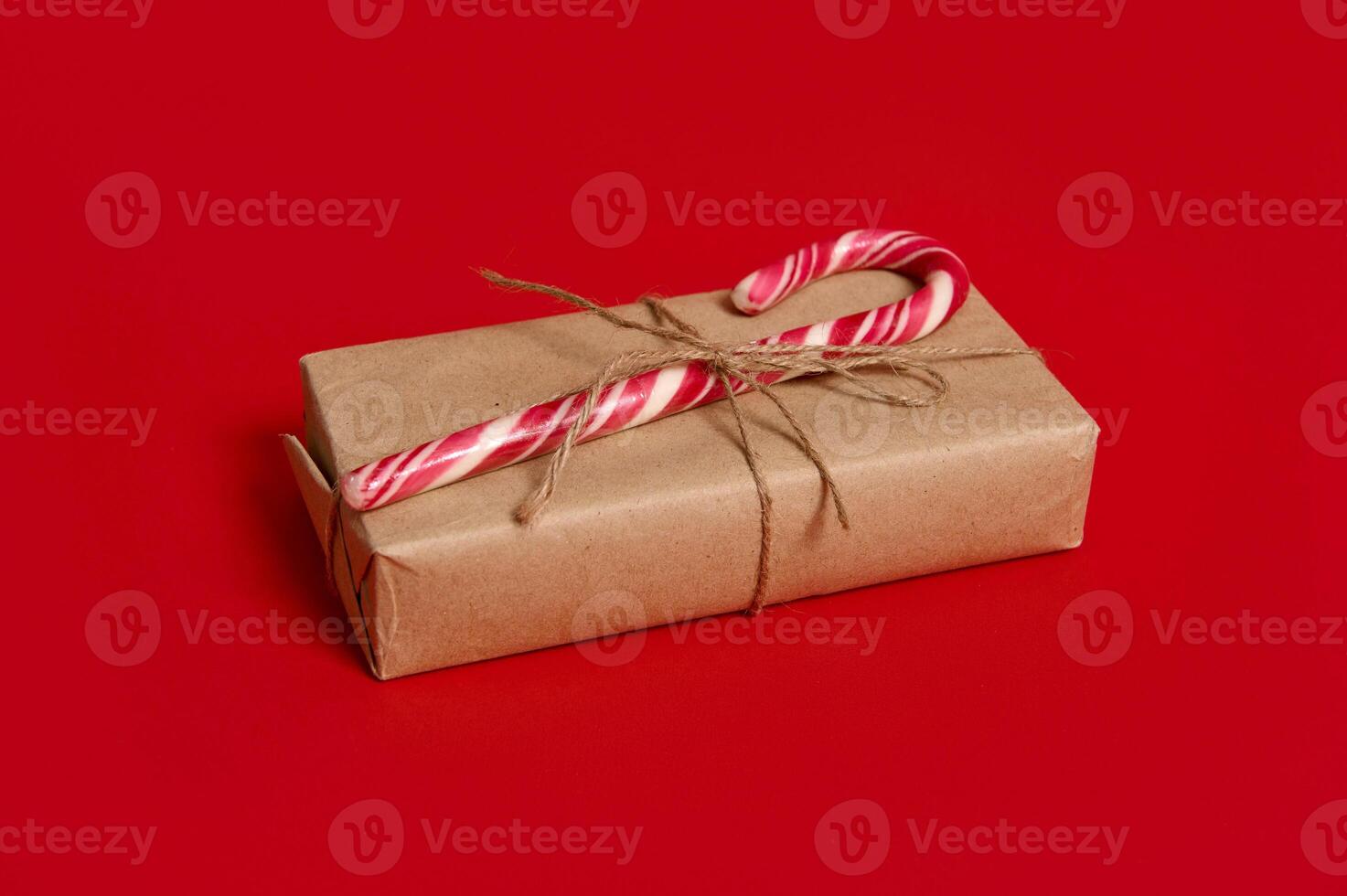 Beautiful minimalist composition of Christmas items, gift box wrapped in kraft wrapping gift paper with tied bow and sweet candy cane isolated over red background with copy space for advertisement photo