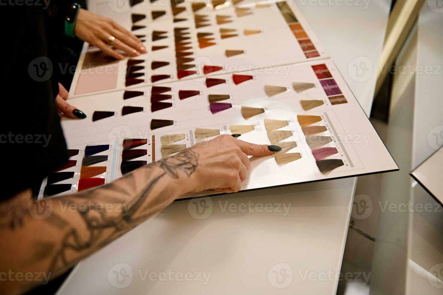 Cropped image of a well-groomed tattooed hairdresser arm pointing to a swatch of straight dyed hair in a hair dye color palette catalog and advising a client on hair coloring in beauty salon. Close-up photo