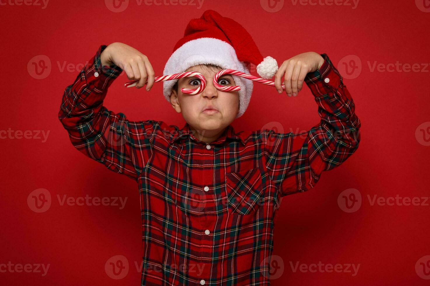 Adorable preteen boy in Santa Claus hat and plaid shirt looks at camera through Christmas sweet candy canes, holding them imitating eyeglasses, posing on red background with copy space for ad photo