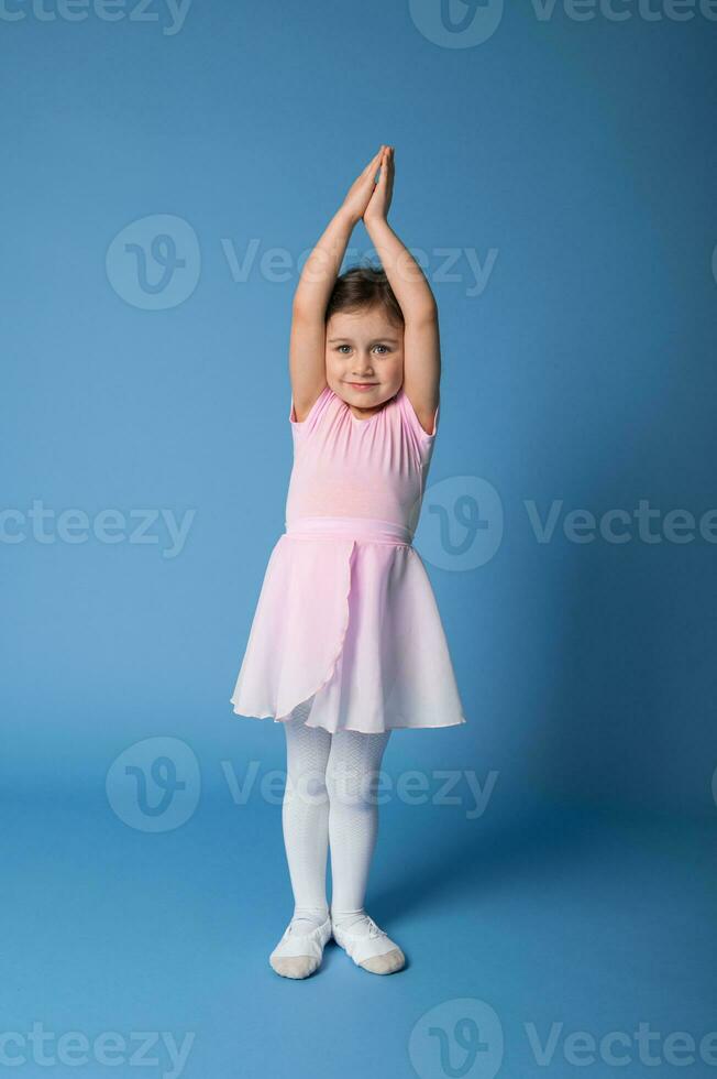 Adorable ballerina in pink dress stands on blue background and raises her arms up thereby stretching her body photo