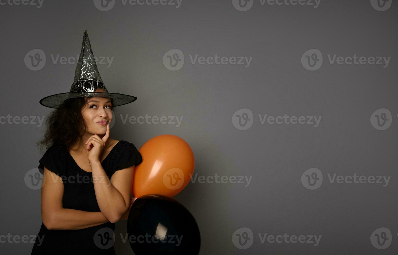 Beautiful pensive mysterious young mixed race woman in black and wizard hat, looking up thoughtfully , posing against a gray background with copy space for ad. Halloween party concept photo