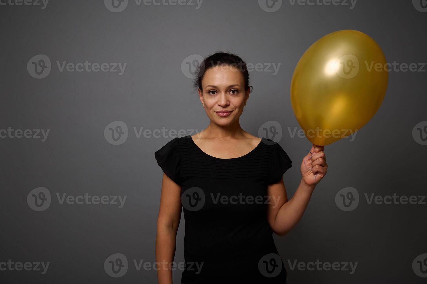 alegre africano americano mujer vestido en negro sonrisas mirando a cámara y poses en contra gris antecedentes con un oro de colores aire globo en mano. negro viernes concepto con Copiar espacio para anuncio foto