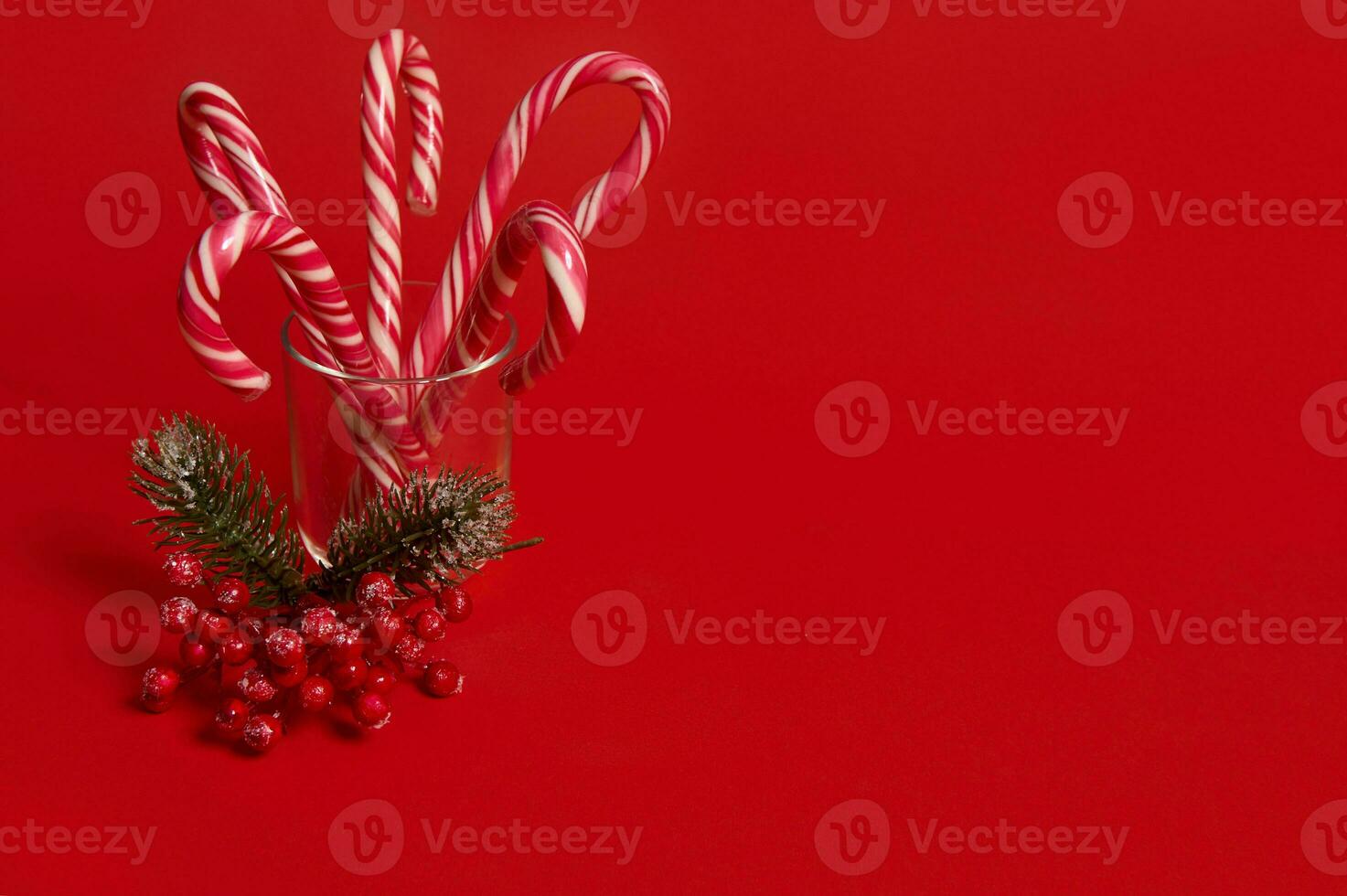 estudio Disparo de hermosa minimalista sencillo composición con Navidad piruletas en transparente vaso y Nevado rama de pino con rojo bayas, acebo, en rojo antecedentes con Copiar espacio para anuncio foto