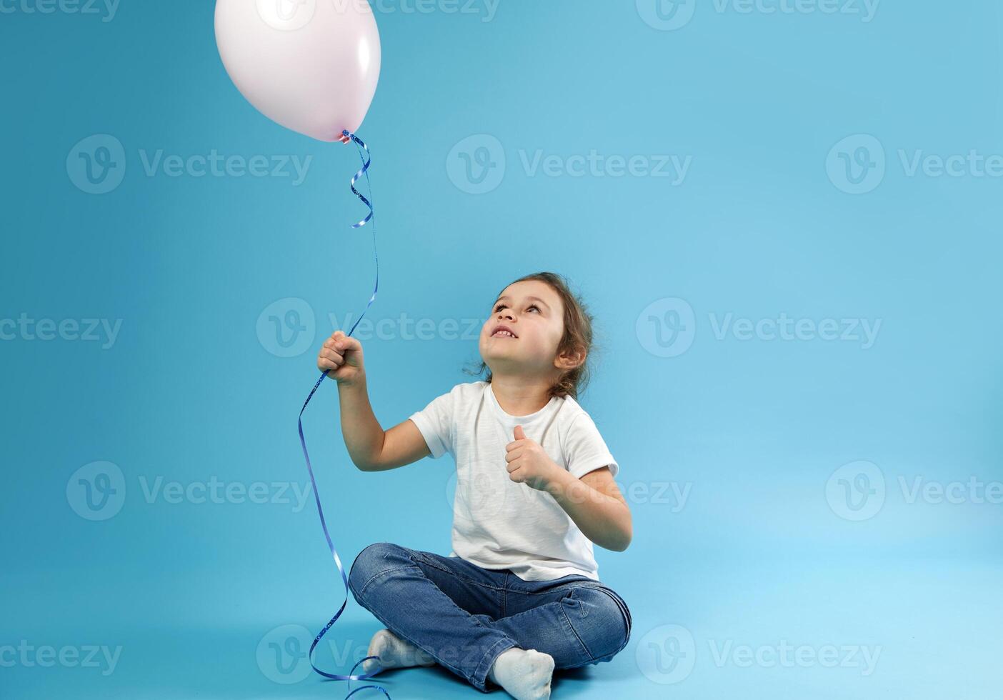 Smiling little girl sitting on blue background with soft shadow and holding balloons in the hand. Childhood and child protection day concept. Copy space photo