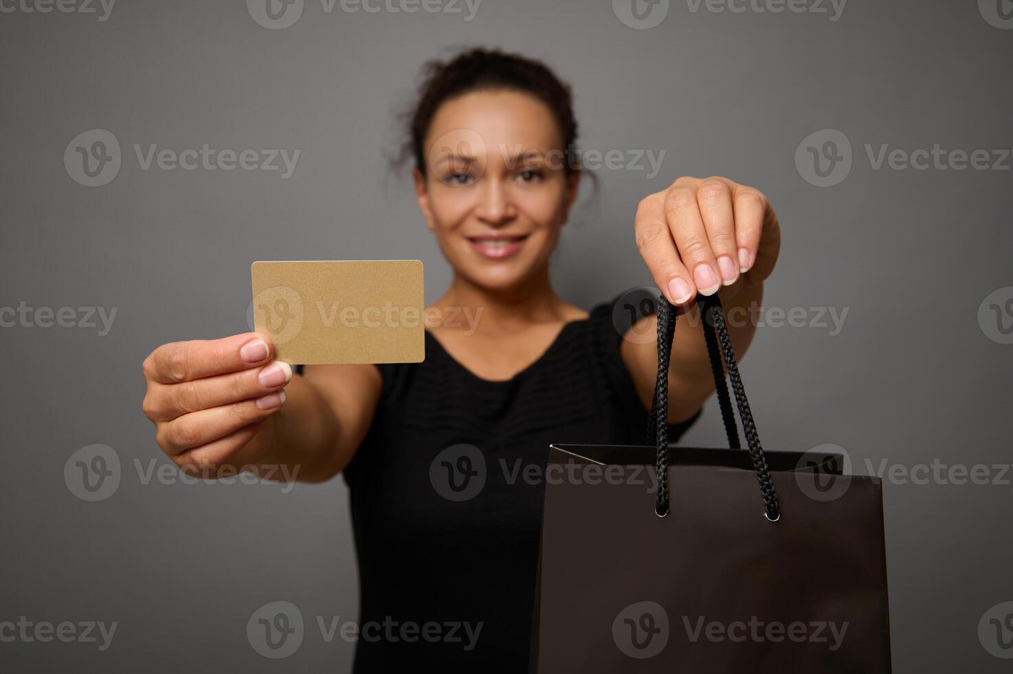 Focus on blank gold plastic credit or discount card and black shopping bag in the hands of blurred pretty woman with beautiful smile, isolated over gray background with copy space for Black Friday ad photo