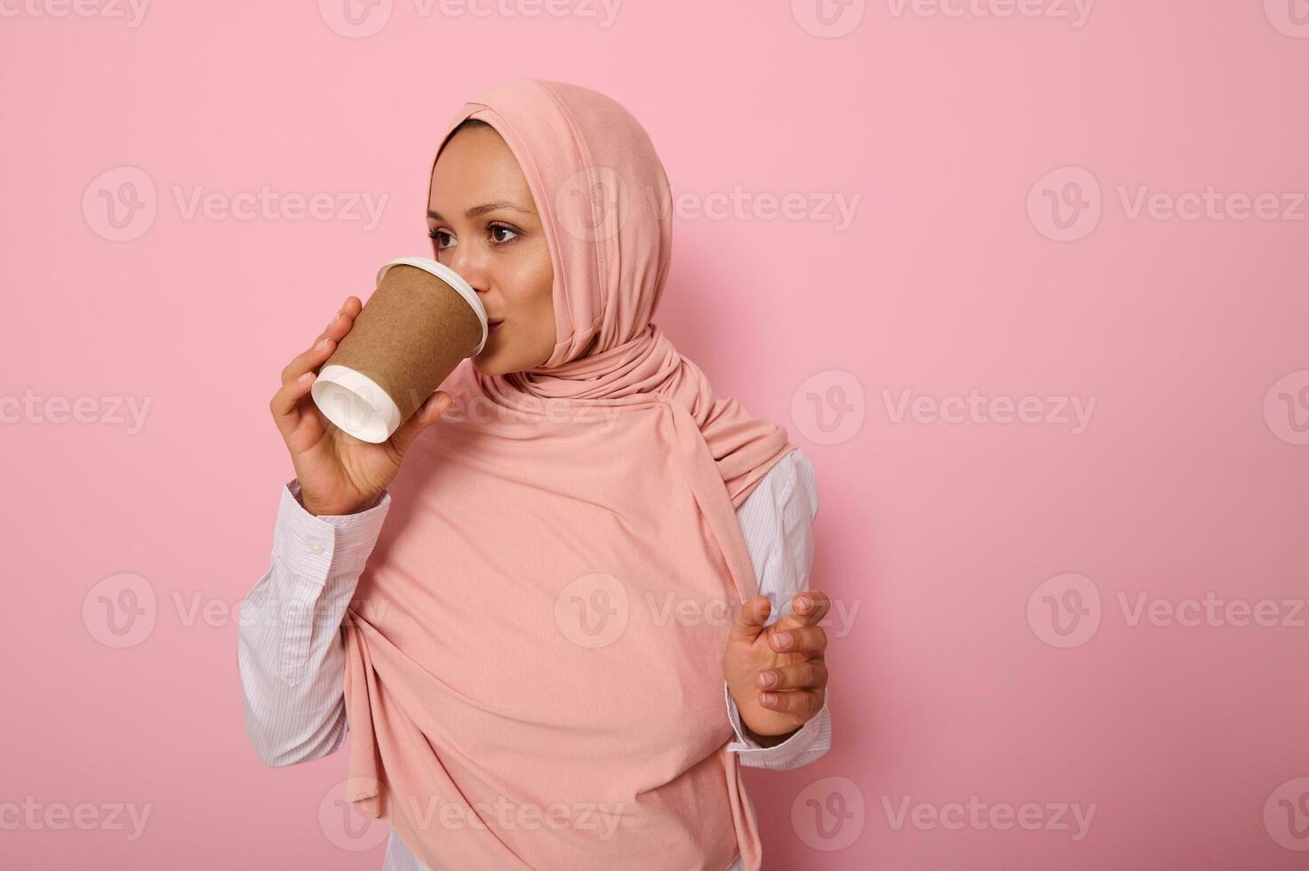 Beautiful Arabic muslim woman with covered head drinking hot drink, tea or coffee from disposable cardboard takeaway cup, standing sideways, three quarters, against pink background with copy space photo