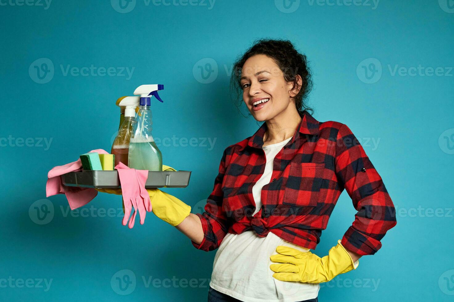 sonriente mujer posando en azul antecedentes con bandeja con limpieza productos en manos y uno mano en cintura foto