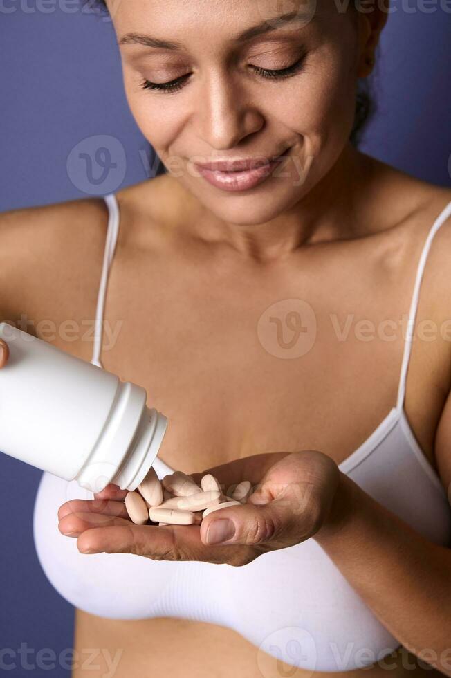 Close-up portrait of a charming young woman in white underwear holding a bottle of diet supplement pills and pouring a vitamin complex into her hand. Medicine, health, skin and body care concept photo