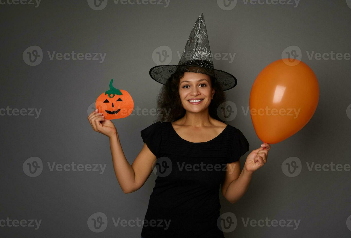 Cheerful woman in wizard hat, dressed in black , holds a colorful orange air balloons and shows a felt-cut handmade pumpkin, smiles with beautiful toothy smile looking at camera. Halloween concept photo