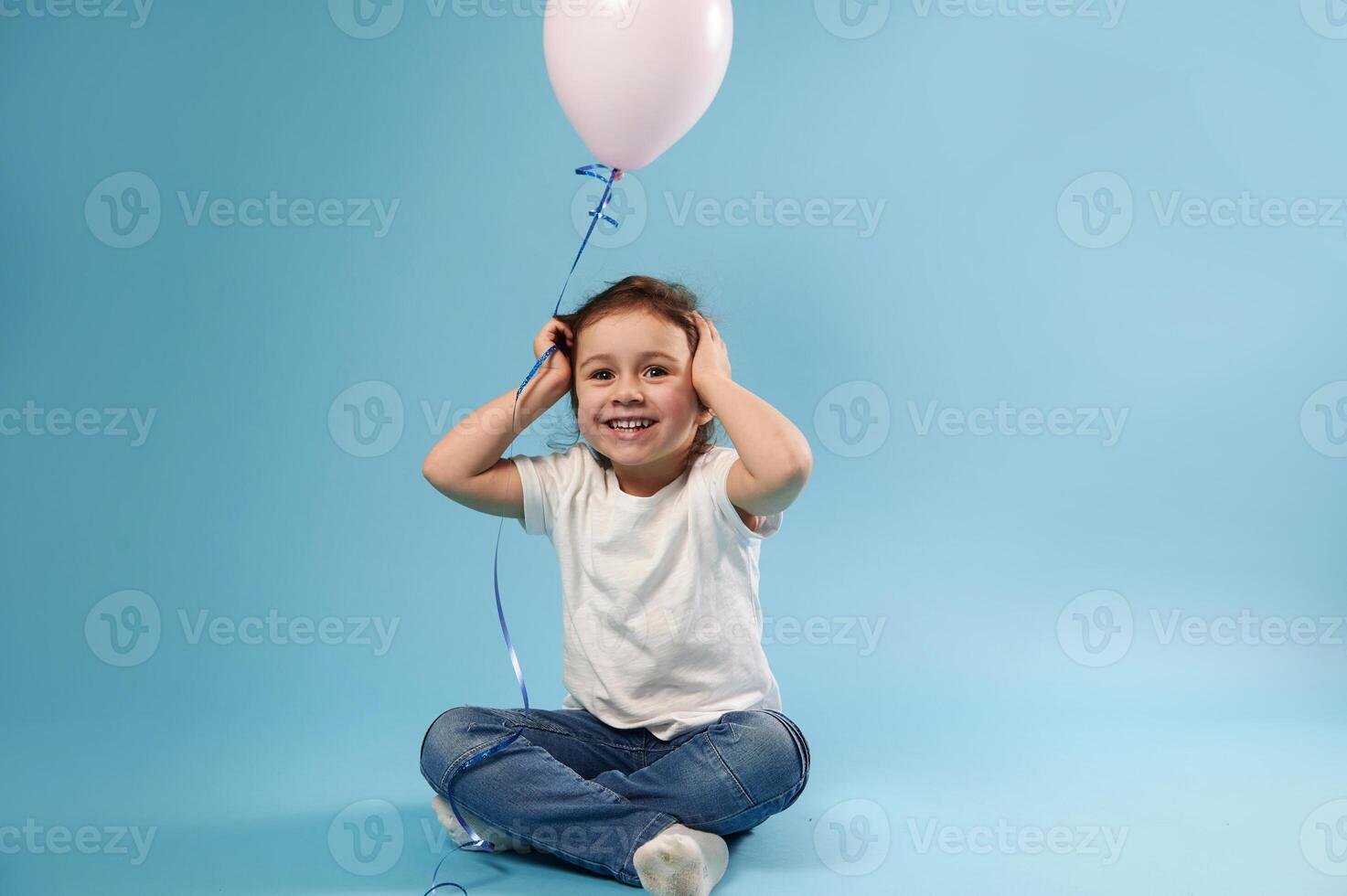 Joyful girl in white t-shirt and blue jeans sits on blue background, puts her hands on her temples and cute smiles looking at camera expressing happiness. photo