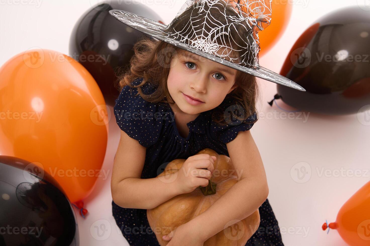 4 years old adorable pretty little girl in witch dress and wizard hat looks at camera playing with balloons and a pumpkin isolated on white background. Concept of a child having fun at Halloween party photo