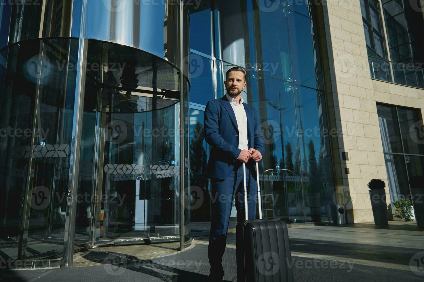 Handsome successful confident businessman in a business suit with black luggage against the background of a glass entrance to a modern corporate high-rise building photo