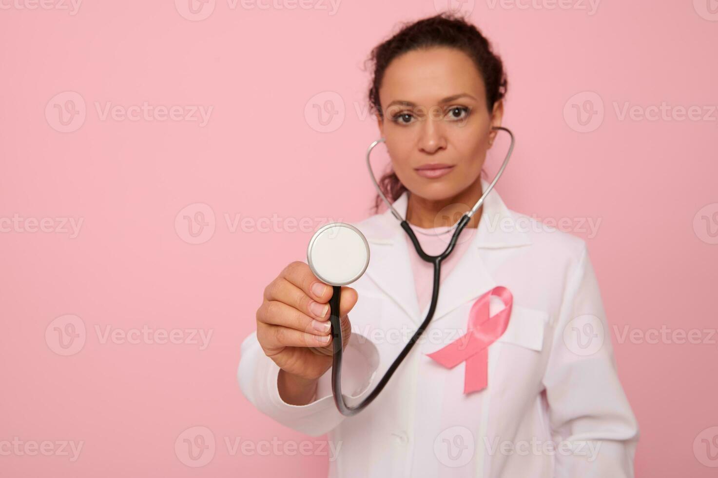 Focus on phonendoscope in the hands of beautiful mixed race Hispanic doctor in medical coat with pink satin ribbon, symbol of Global Breast Cancer awareness Day, 1 st October . Woman's health concept photo