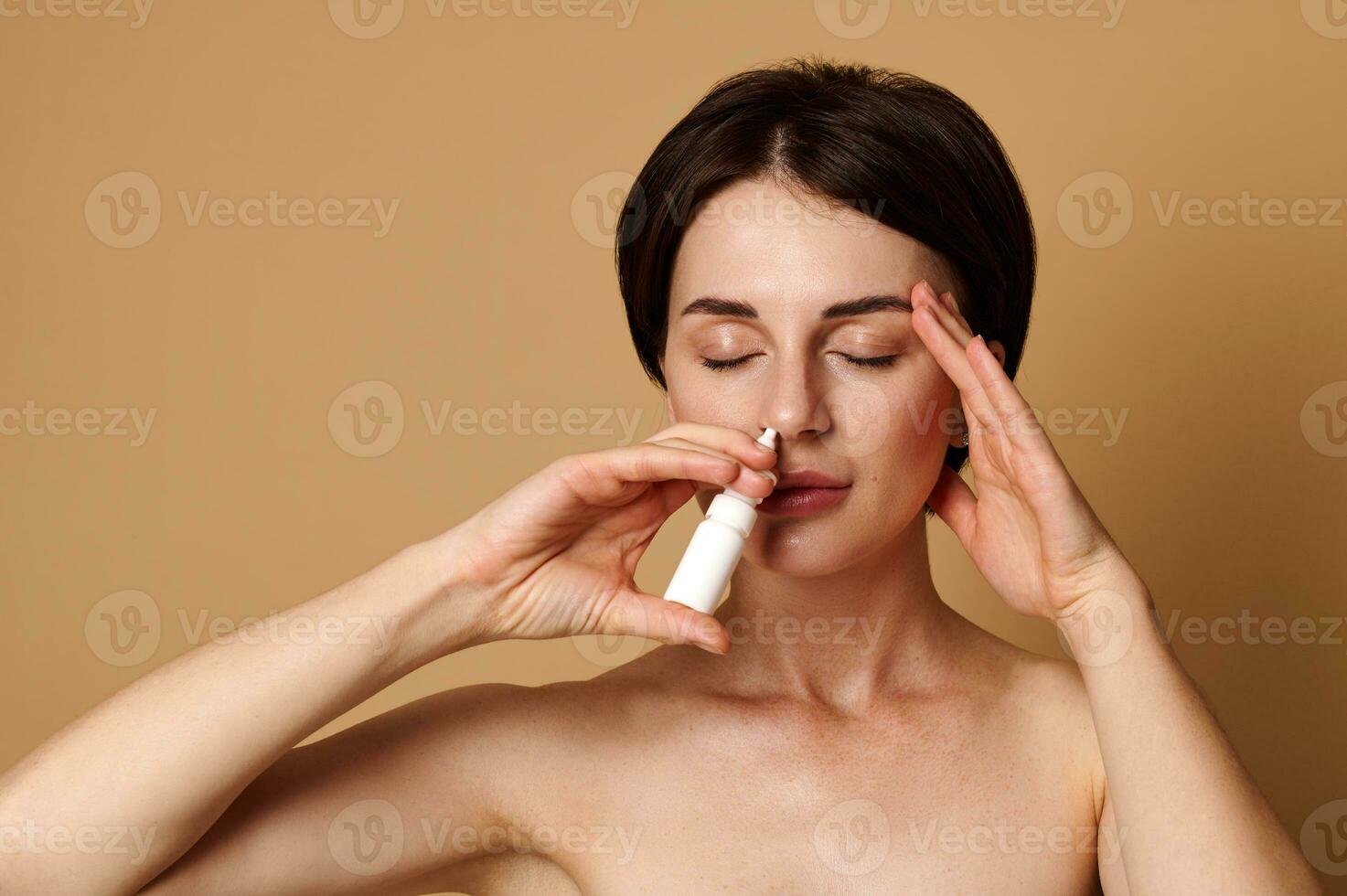 The treatment of rhinitis. Young sick European girl holding a nasal spray and dripping nose drops, isolated over beige background with copy space for medical advertisement photo