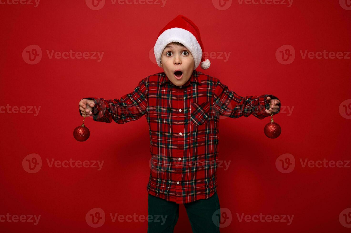 Adorable amazed preteen boy, surprised beautiful child stands on a red background with Christmas spherical shiny toys in his arms, looks at the camera with surprise. New year concept with copy space photo