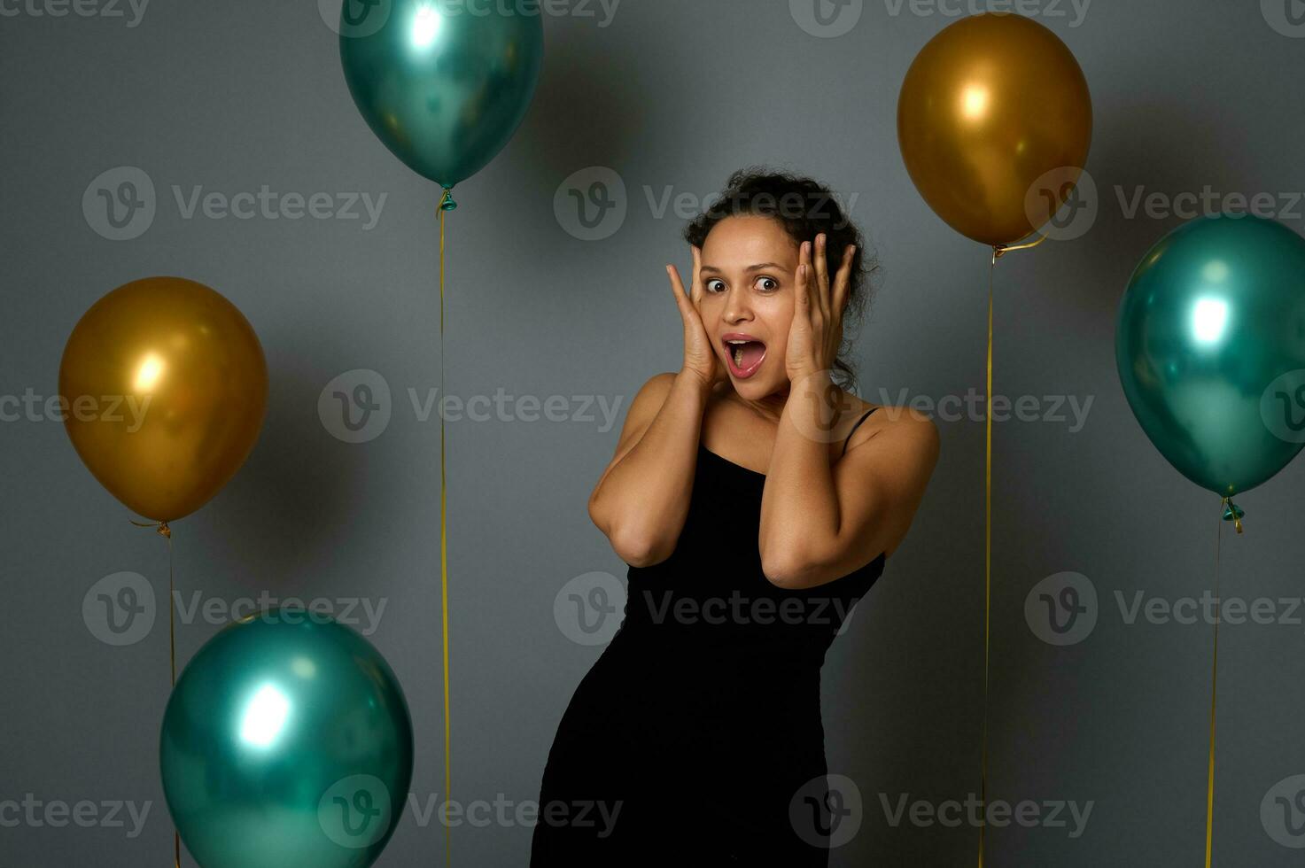 Portrait on gray wall background of an amazed Hispanic pretty woman in black evening dress looking at camera with hands on face, against gray background with luxury air balloons. Celebration concept photo