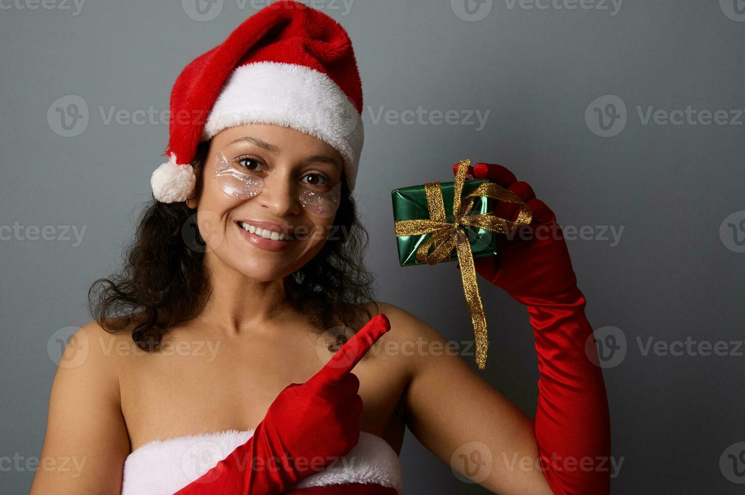Cheerful African woman wearing cosmetic patches under eyes, dressed as Santa points with finger on Christmas gift box in shiny green wrapping paper and golden bow, smiles looking at camera. Copy space photo