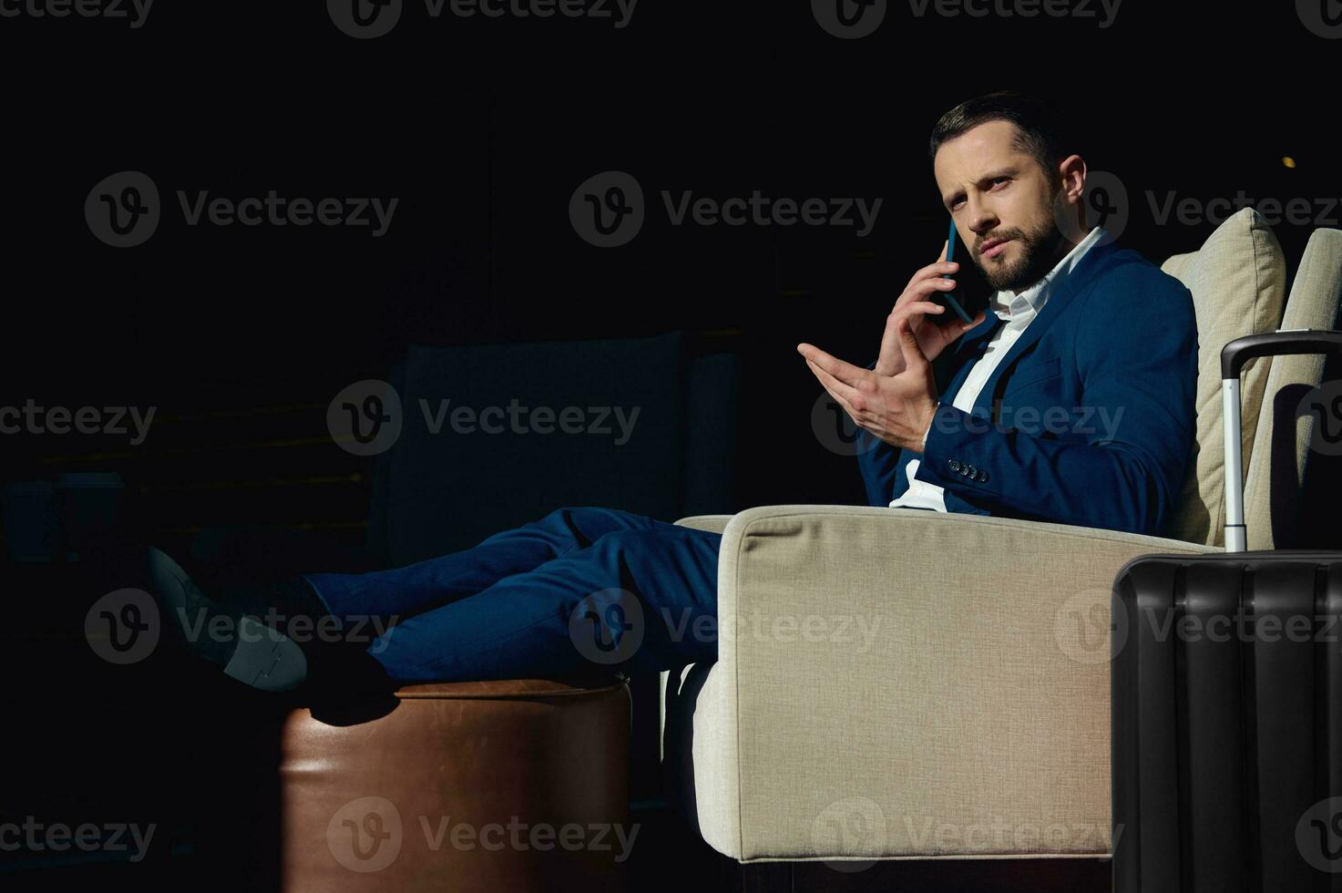 Confident successful and perplexed young European businessman talking on a mobile phone while sitting in an armchair of a hotel lounge, relaxing after a business trip. Suitcase near armchair photo