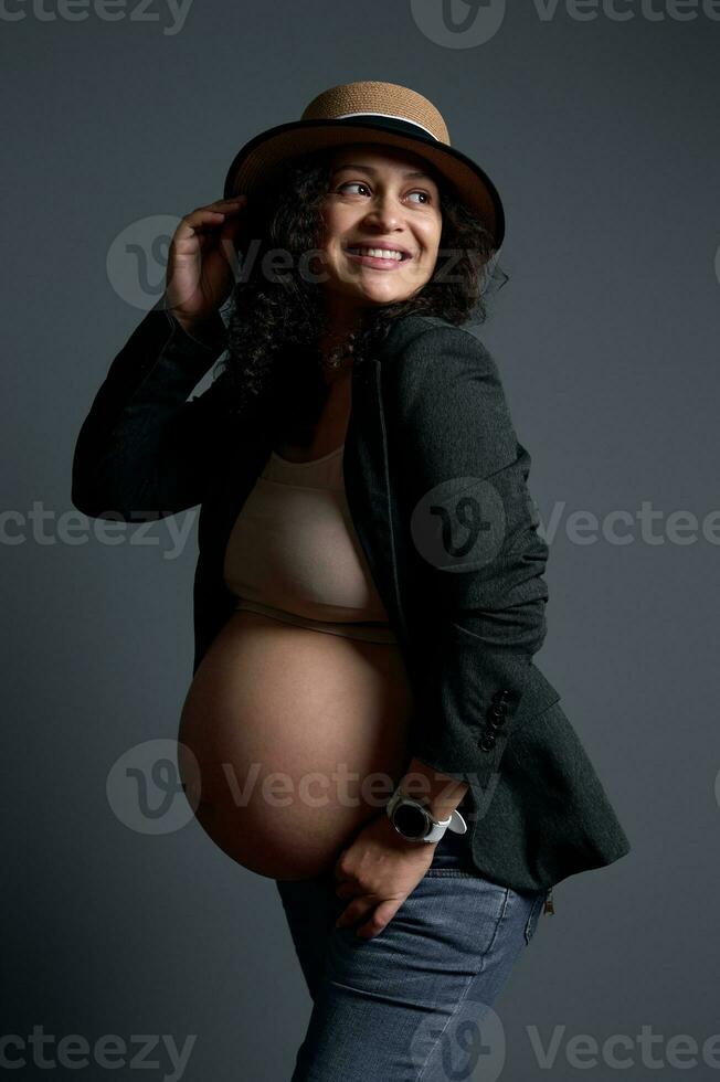 Stunning pregnant woman in straw hat and stylish blazer over lingerie, smiling broadly, looking aside a copy ad space photo