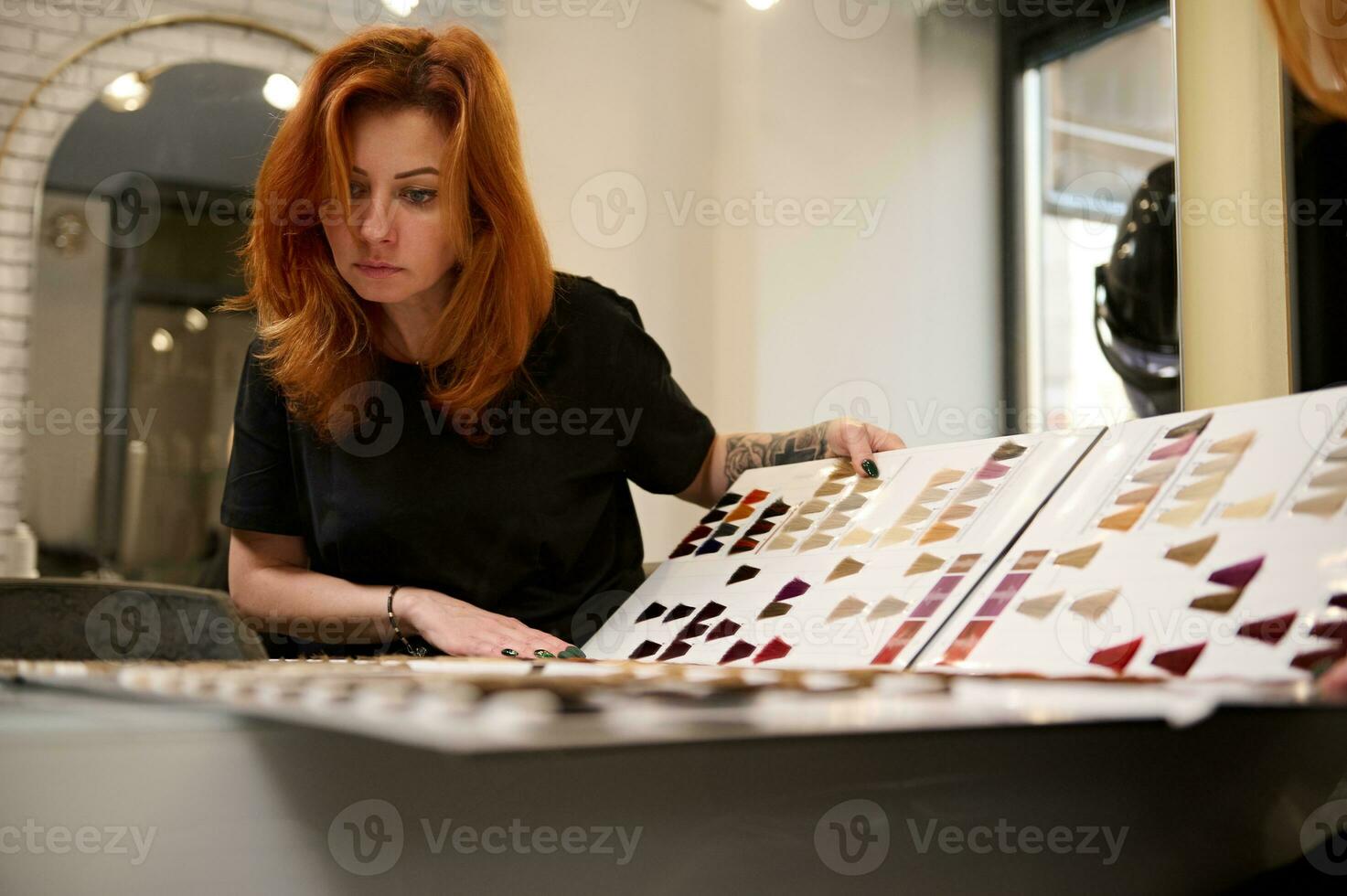Reflection in the mirror of a concentrated stylish beautiful redhead woman with tattooed arms, professional hairdresser with a palette of different colors to hair dye at hairdressing salon. photo