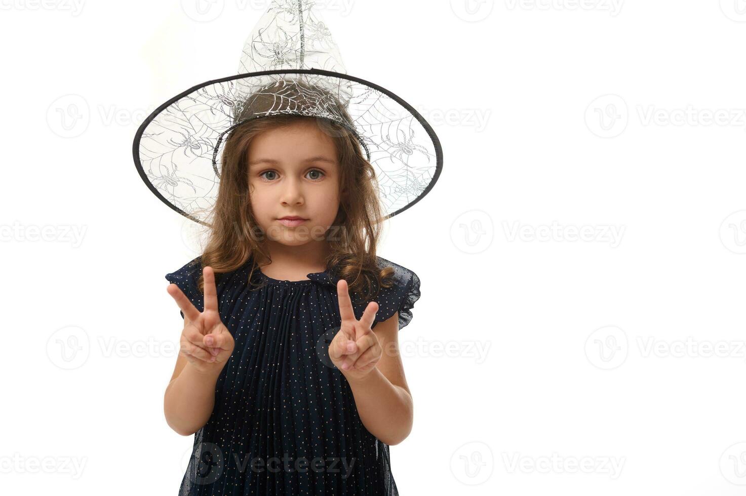 aislado cintura longitud retrato de bonito pequeño bruja niña vistiendo un mago sombrero y vestido en elegante carnaval vestido, gesticulando, demostración un paz firmar con dedos. Víspera de Todos los Santos concepto foto