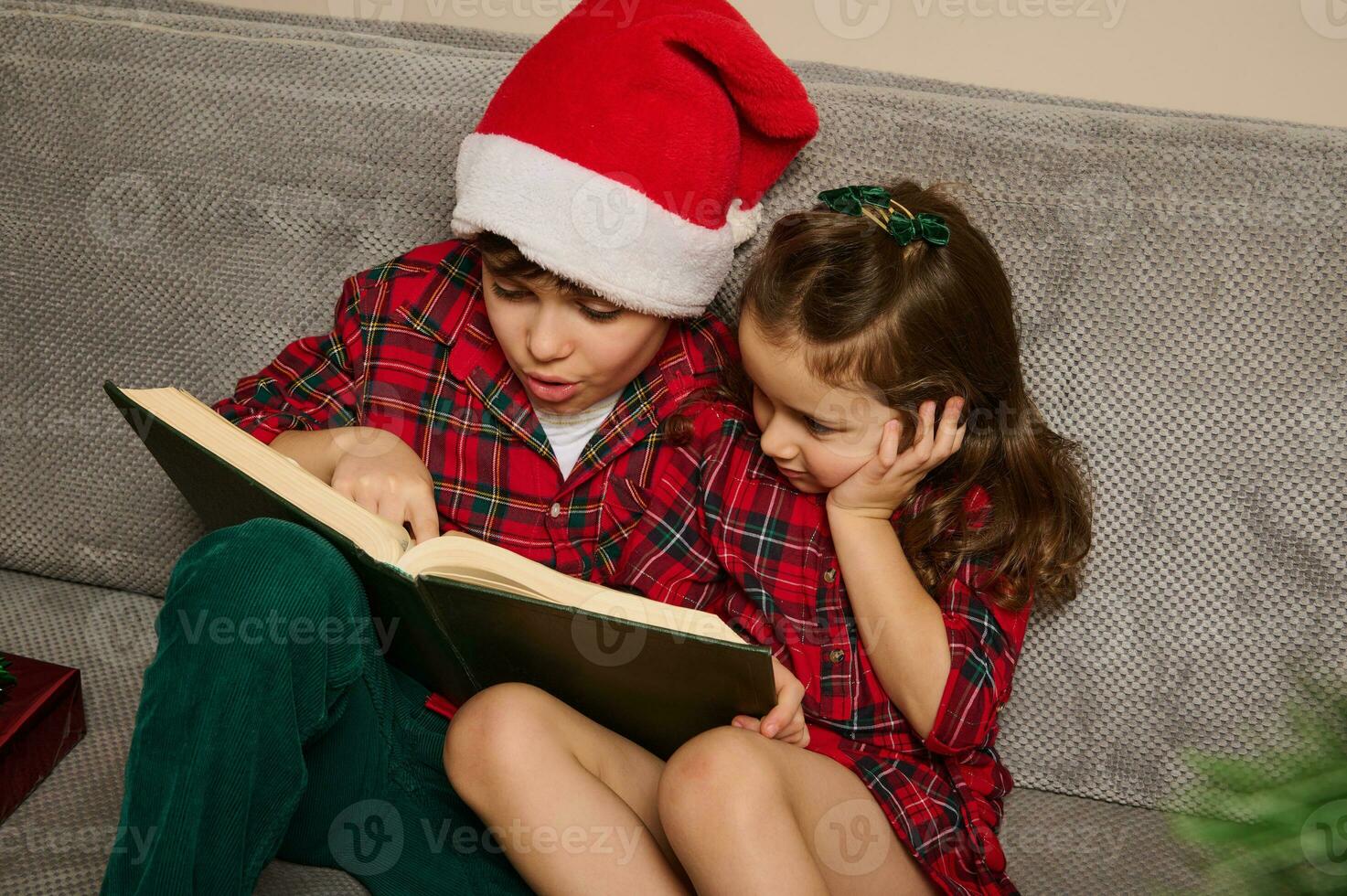 Adorable Caucasian children , brother in Santa's hat and his younger sister sitting on the couch and reading a book, fairy tales on the Christmas night at home photo