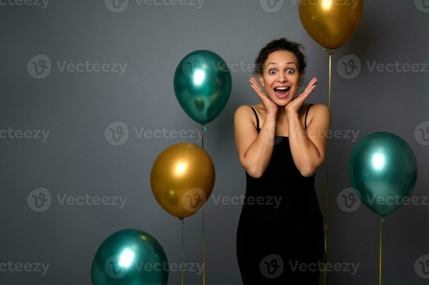 Beautiful woman in black dress holds her hands on her cheeks, poses with opened mouth expressing happiness, surprise. Birthday, Christmas and events concepts on gray background with copy space for ad photo