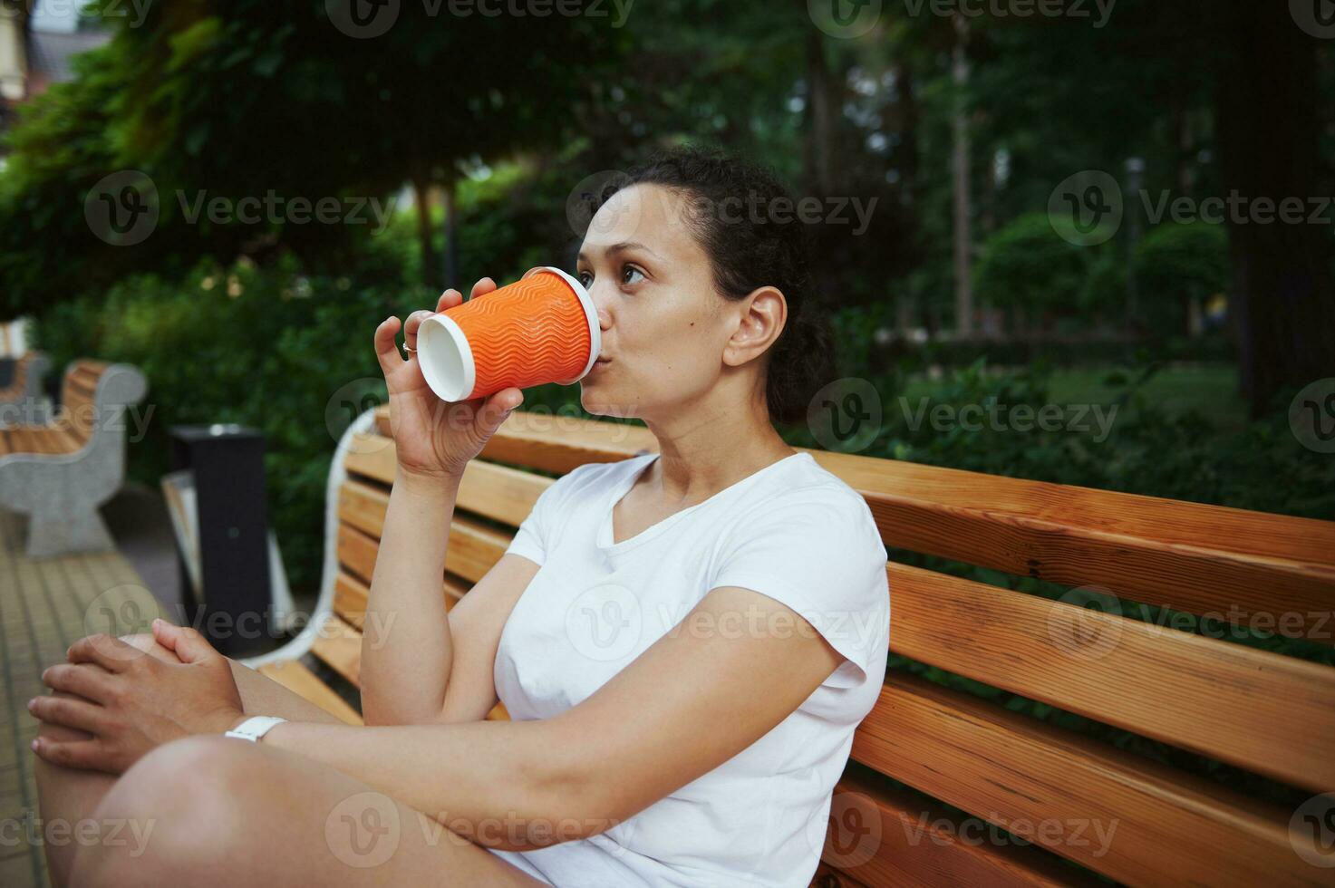 joven multiétnico mujer Bebiendo para llevar café desde un desechable papel taza, sentado en de madera banco en un verano parque foto