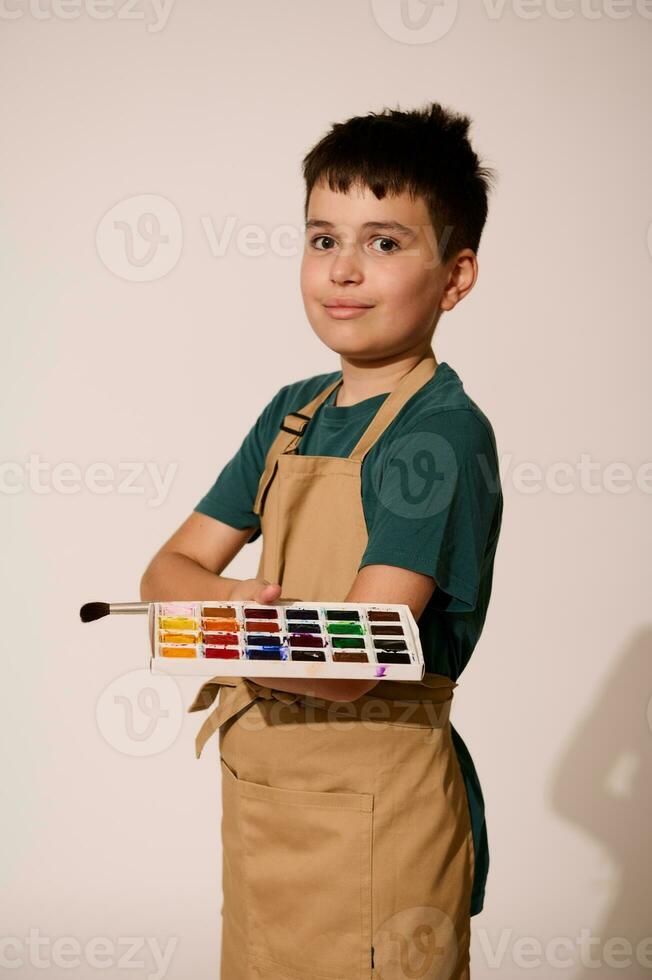 Teenage boy in beige apron, holds paintbrush and palette with watercolors, smiles at camera, isolated studio background photo