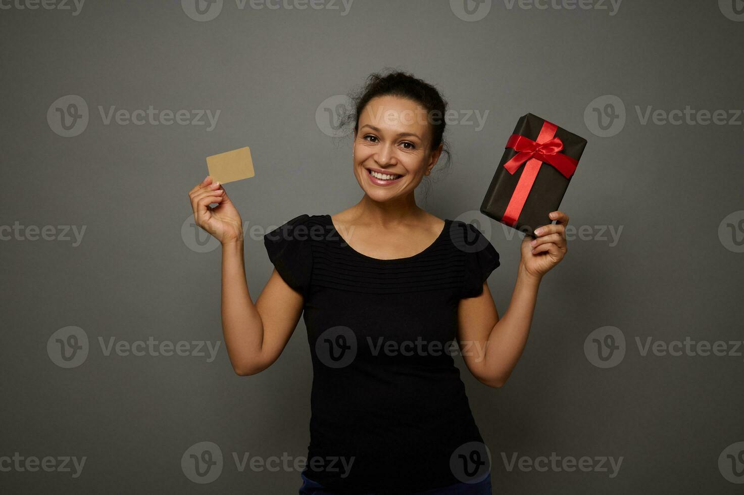 Mixed race beautiful woman dressed in black holds a present in black wrapping gift paper and a gold credit card and shows them smiling to camera, isolated over gray wall background with copy space photo