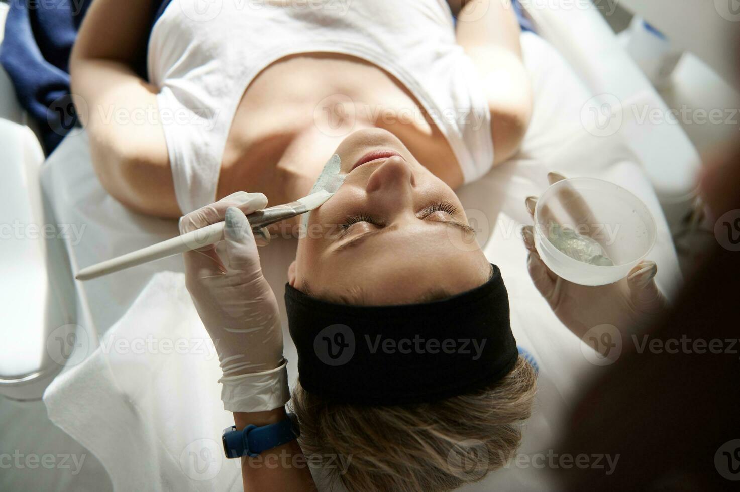 Overhead view of young woman relaxing on a massage table during beauty spa procedure and doctor cosmetologist applying alginate mask on her face photo