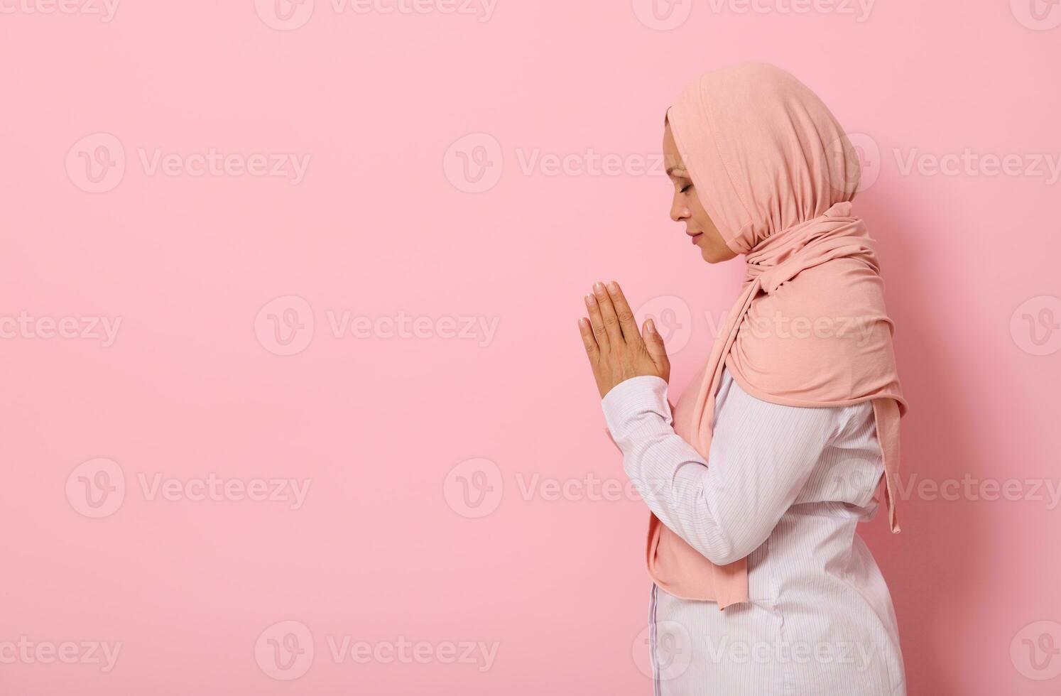 Side portrait of a beautiful and serene Muslim Arab woman in pink hijab and strict outfit with palms folded together at face level performing namaz, isolated on colored background with space for text photo