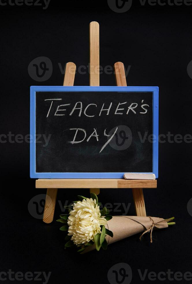 Close-up of a bouquet of asters flowers next to a chalk chalkboard with lettering Teachers Day, standing on a wooden table easel, isolated on black background with copy space. photo