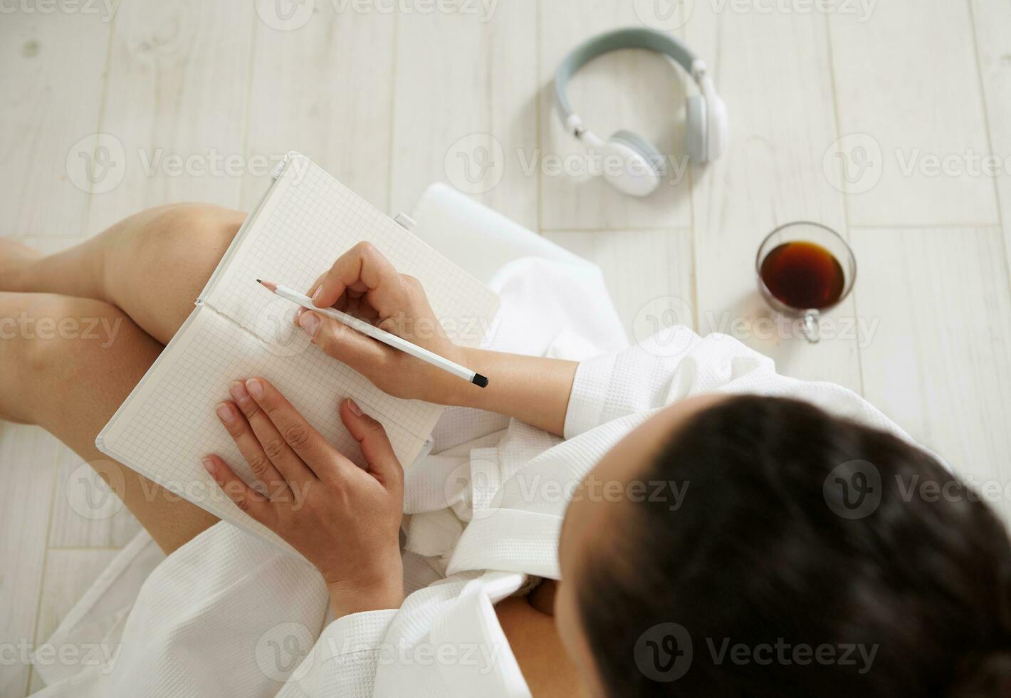 parte superior ver de un morena mujer sentado en el piso y escritura en un cuaderno. taza con café y auriculares mentira en el piso foto