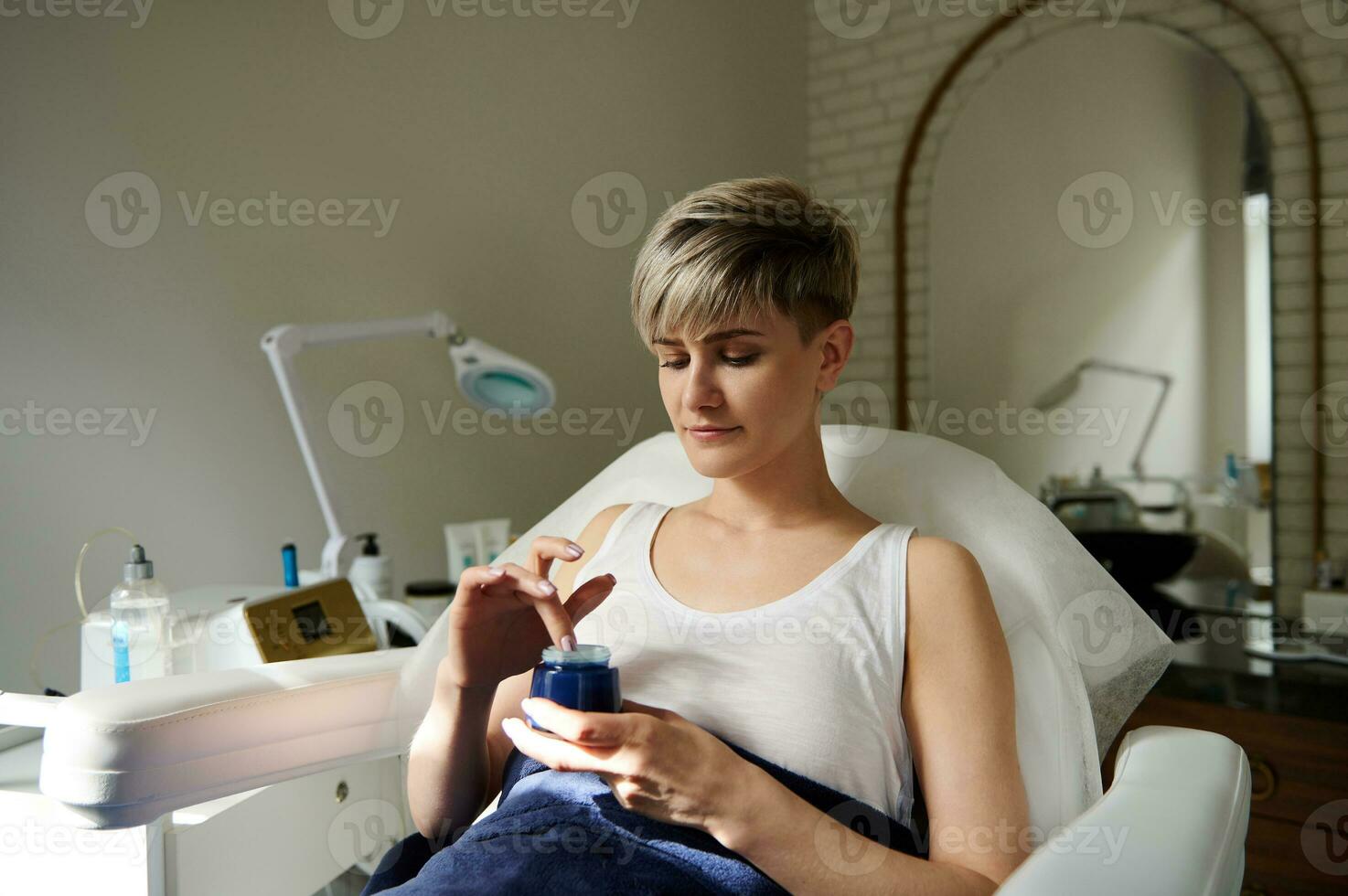 hermosa joven mujer sentado en un silla a cosmetología habitación, participación un botella con hidratante y aplicando eso en cara para piel cuidando foto