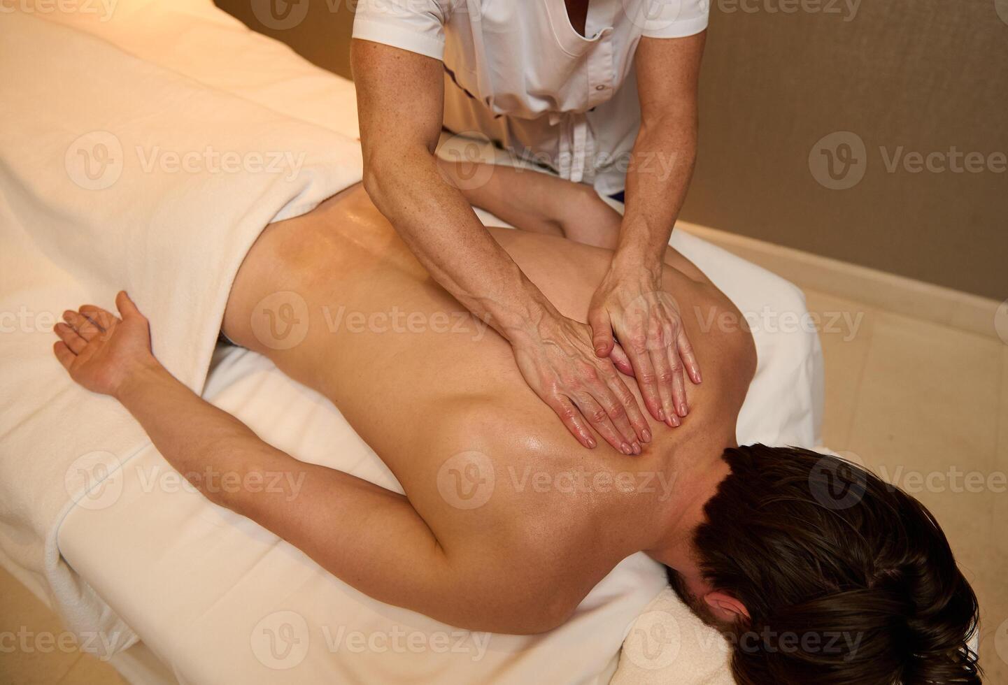 Overhead view of unrecognizable professional massage therapist massaging back muscles of a young man, relaxing, lying down on massage table at luxury wellness spa center. Alternative therapy concept photo