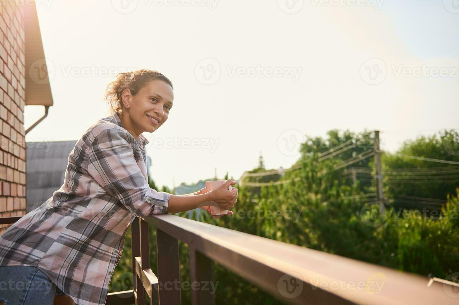 Charming woman drinks coffee, enjoys the view of country houses from balcony on sunny day photo