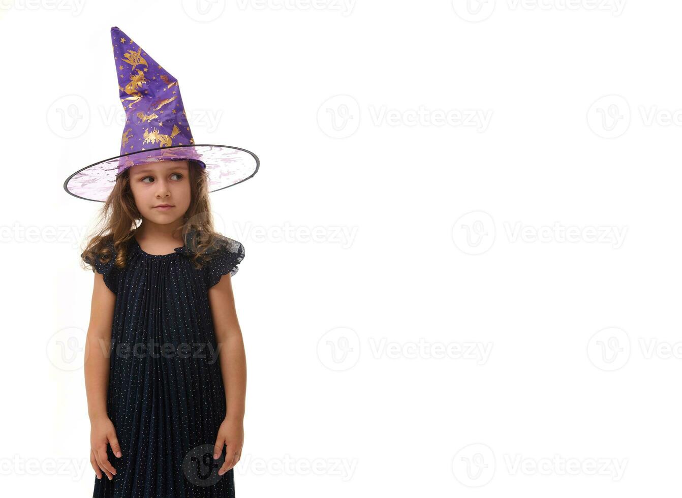 Portrait of pretty little girl wearing a wizard hat and dressed in stylish carnival dress, looking at camera posing with crossed arms against white background, copy space. Halloween concept photo