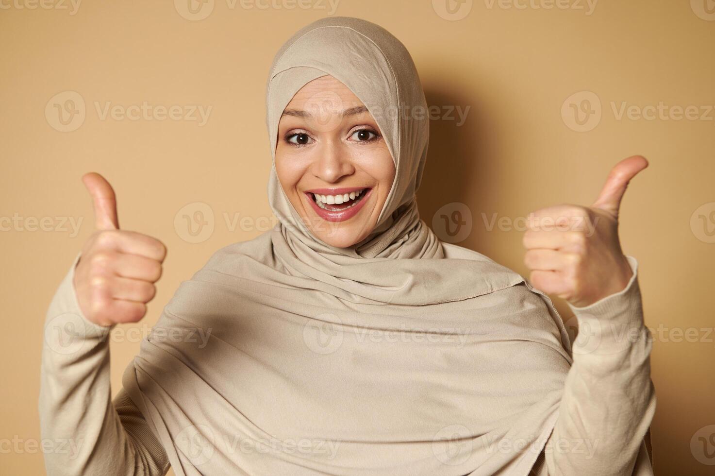Happy Arab muslim woman in hijab with a beautiful toothy smile showing thumbs up to camera. Beige background with copy space. photo
