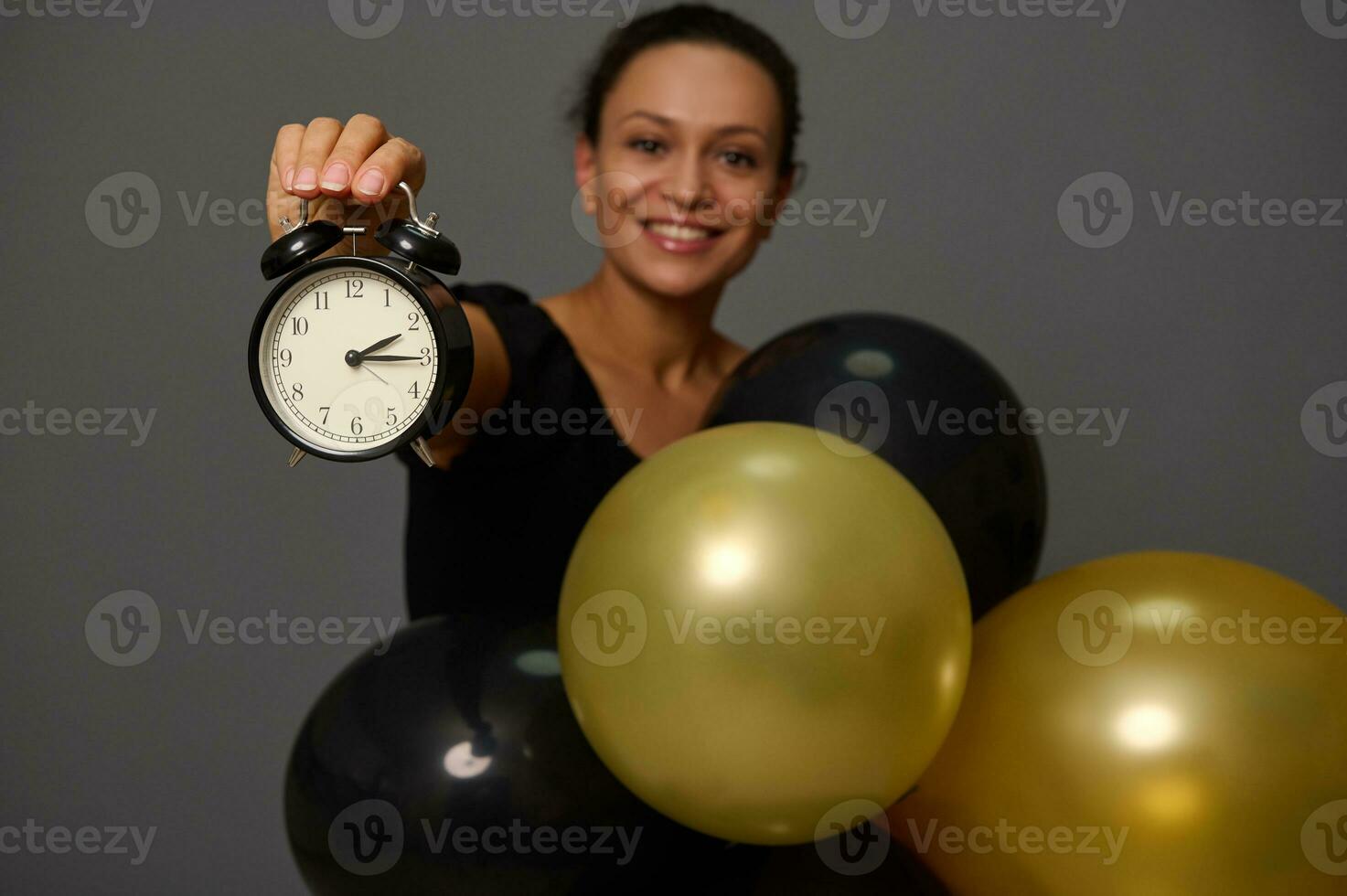 Focus on the hand holding a black alarm clock. Blurred smiling woman with gold black ir balloons gray background with copy space for advertisement Shopping at Black Friday photo