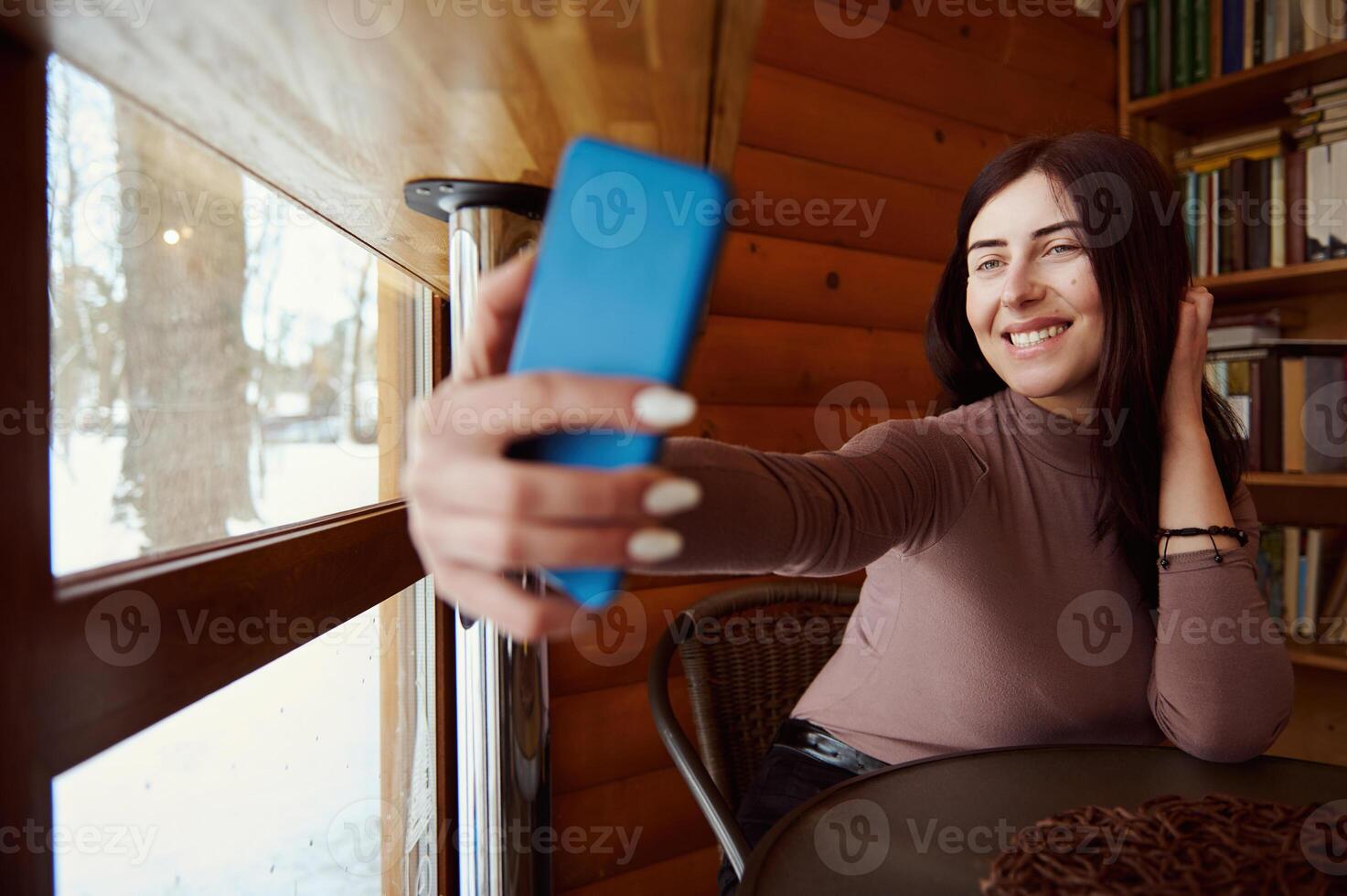 Young attractive woman holds a smartphone and smiles while making selfie sitting atthe cafe on the background of a wooden wall and shelves with books photo