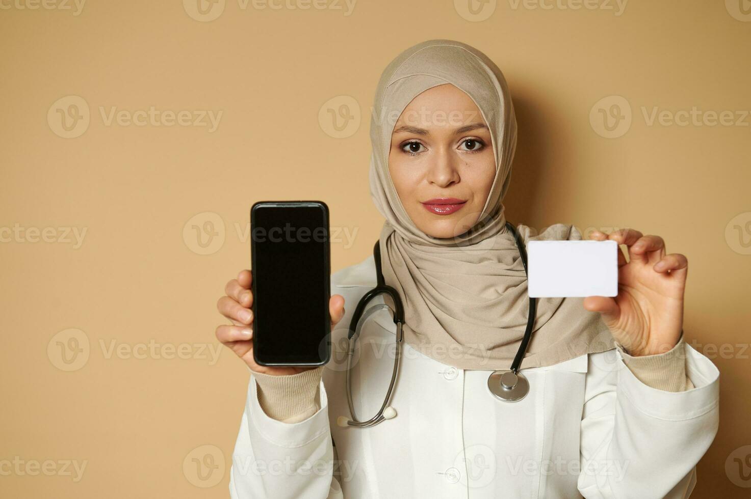 confidente retrato de un joven musulmán hembra médico participación un blanco el plastico blanco tarjeta y teléfono inteligente y demostración ellos a el cámara. seguro concepto foto