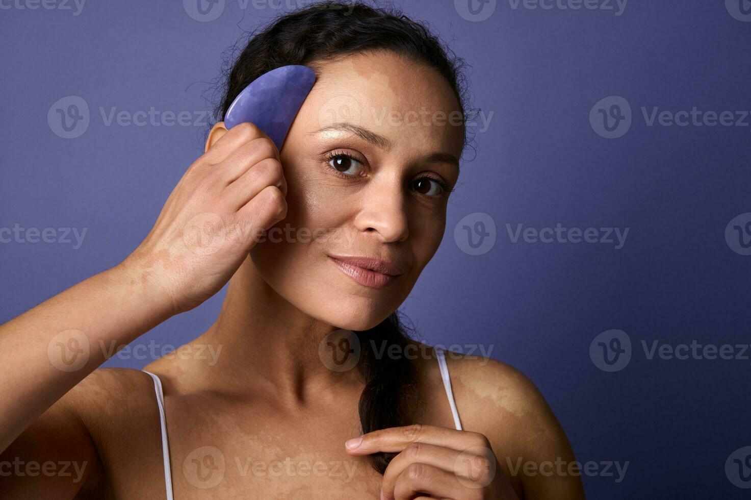 hermosa medio Envejecido mujer con vitiligo sonrisas lindo mirando a cámara, y participación jade Roca masajeador cerca su rostro. púrpura antecedentes con Copiar anuncio espacio. dermatología y piel problemas concepto foto