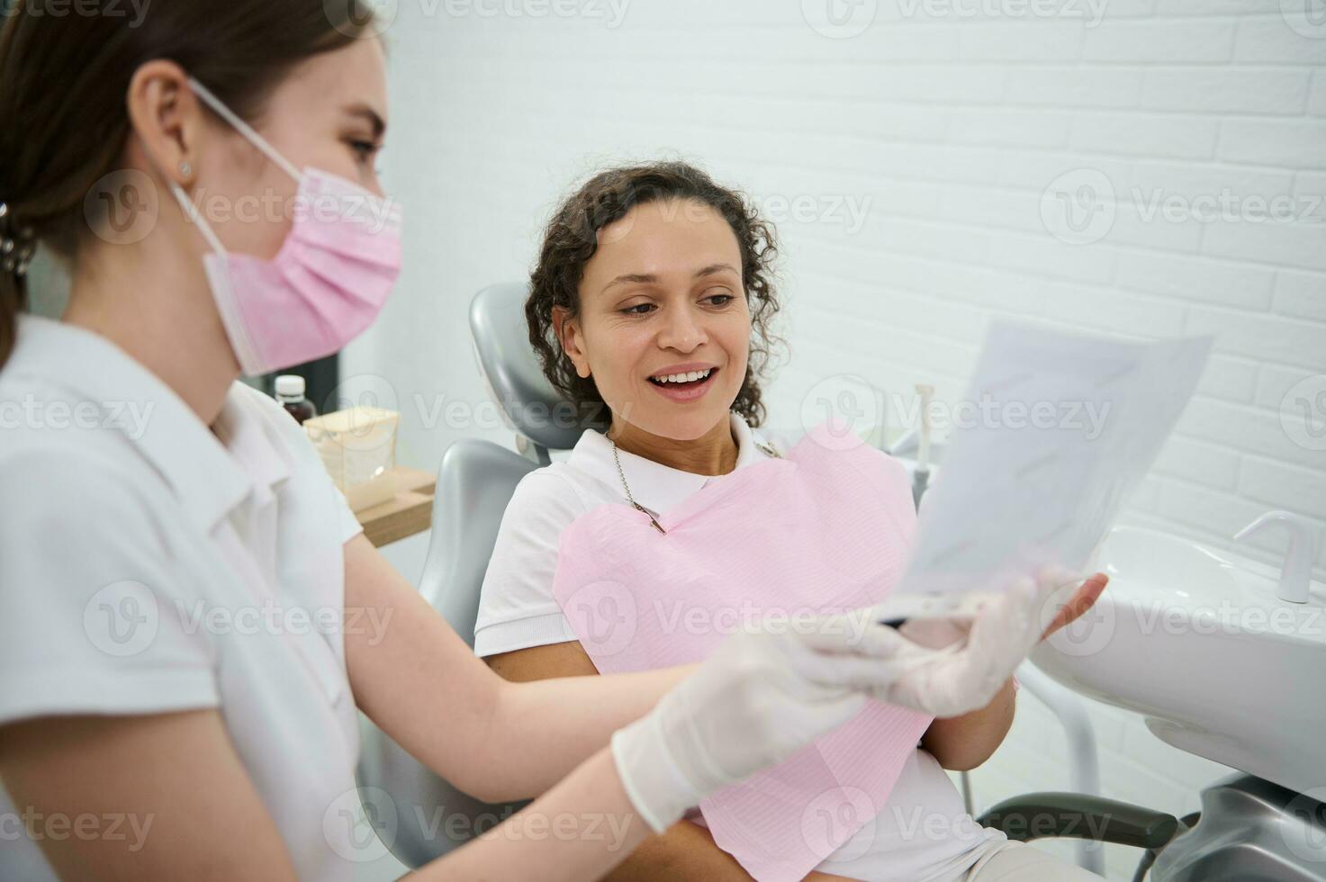 hembra dentista demostración el mandíbula y dientes en un radiografía y que se discute con el paciente durante un dental examen, explicando el consultas tratamiento asuntos, en moderno odontología. foto