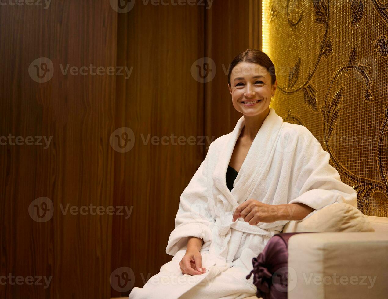 Portrait of a relaxed serene middle aged Caucasian pretty woman in white bathrobe sitting on a couch, smiles toothy smile looking at camera while resting in a luxurious health spa lounge area photo