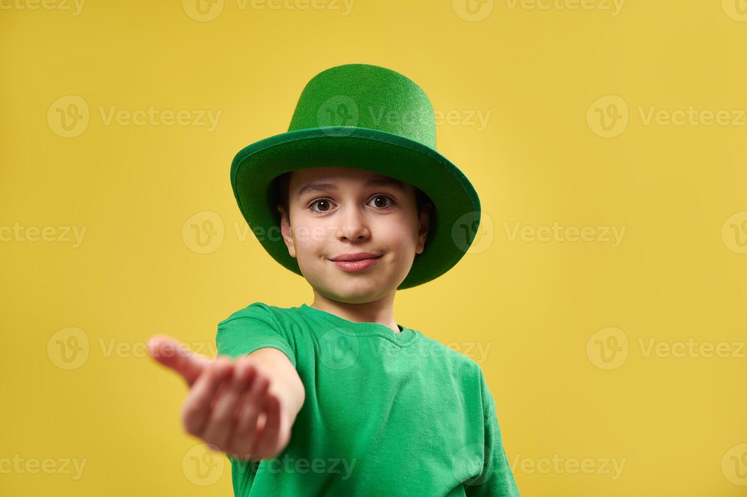 Little boy in leprechaun green Irish hat stretches his palm to the camera. Saint Patrick's Day concept. Copy space and yellow background photo