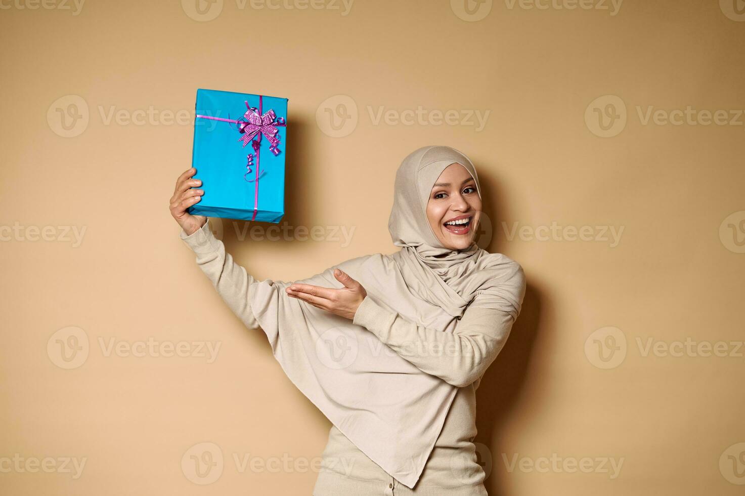 Happy Muslim woman in hijab raising her hands with a gift and presenting it to camera. Beige background, copy space photo