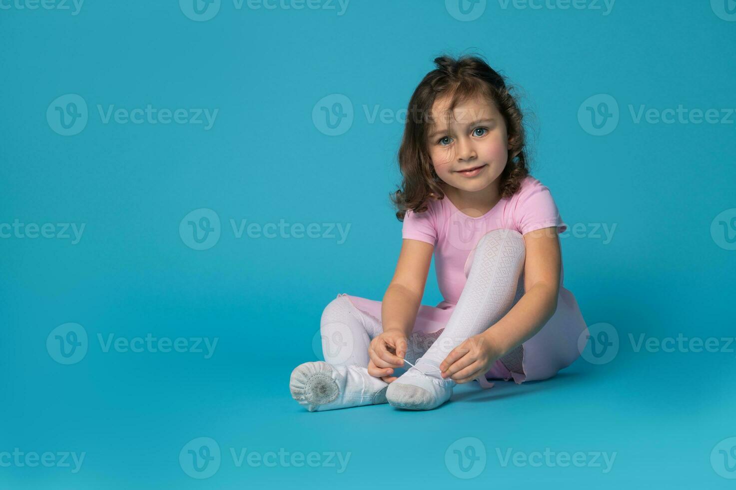 Cute ballerina tying shoelaces on ballet shoes sitting on blue background and looking at camera photo