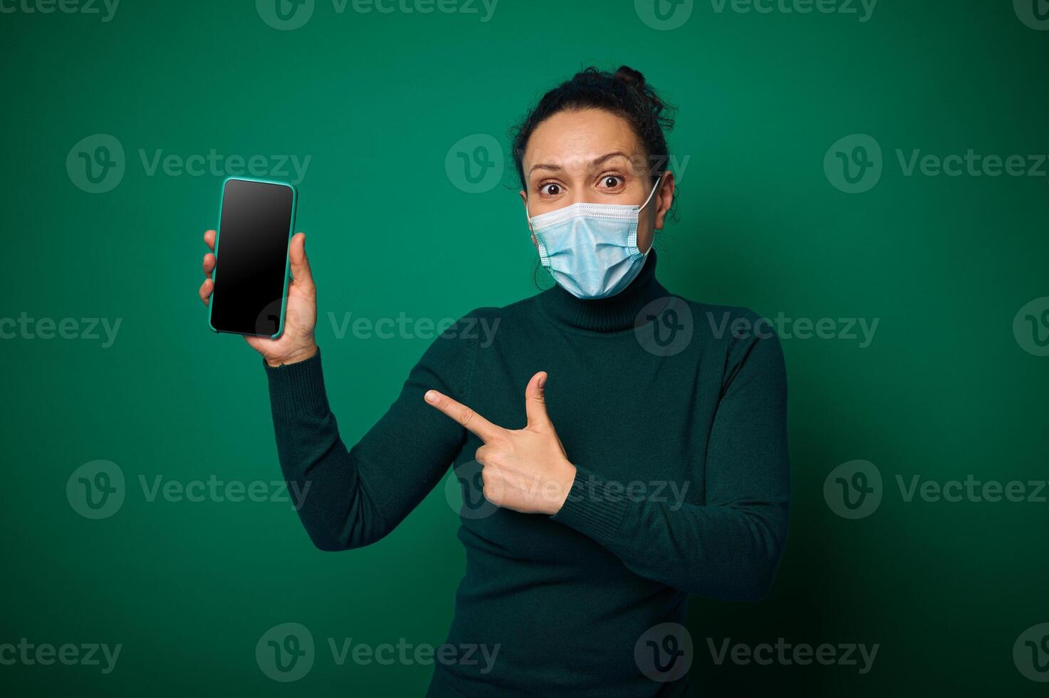 Surprised young woman in protective medical mask looks at camera and points her finger on her smartphone with empty blank screen for copy space, isolated over green background photo